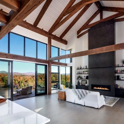 Living room with large windows, exposed wooden beams, fireplace, and scenic outdoor view.