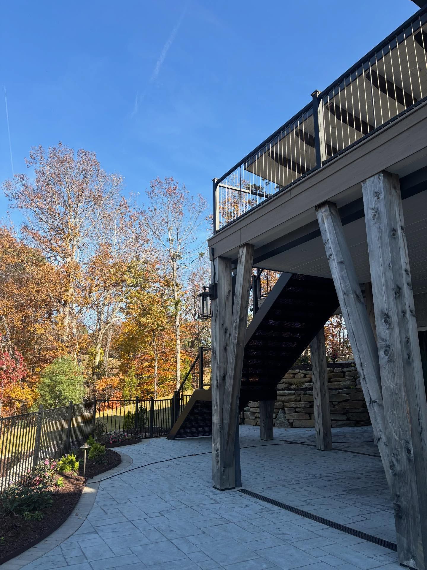 Patio with stone pavers, wooden deck with stairs, trees with fall foliage, and a blue sky.