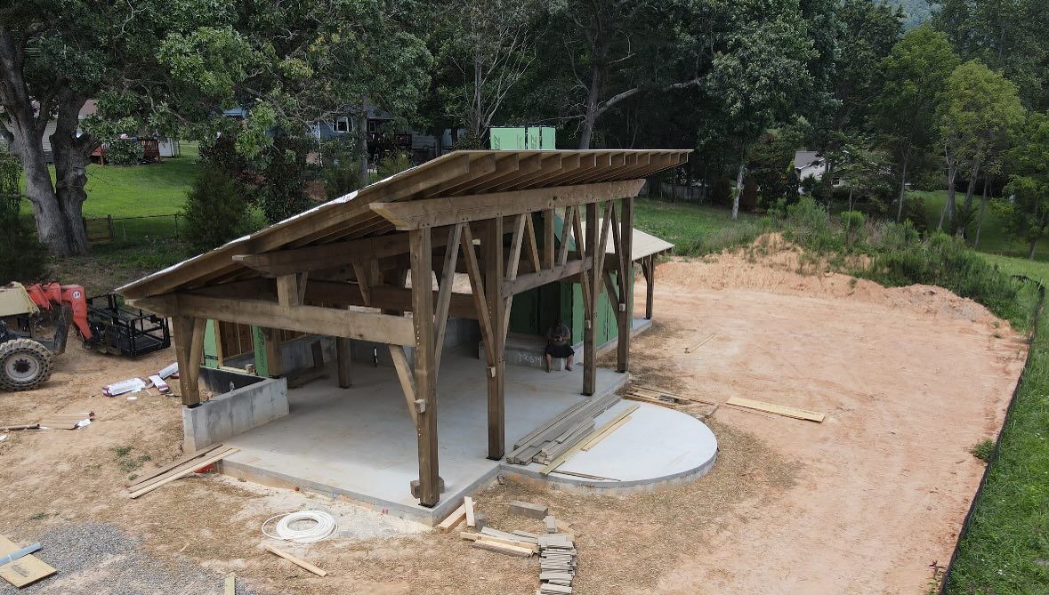 Wooden pavilion under construction on a concrete slab, surrounded by dirt and greenery.