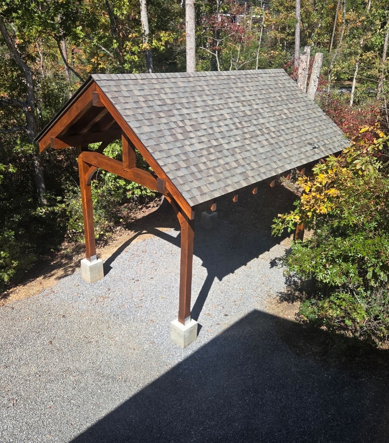 Carport with a shingled roof, supported by wooden posts set in concrete, on a gravel driveway.
