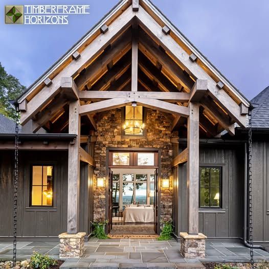Stone and timber entrance of a house, dark gray siding, lit doorway with view, Timberframe Horizons logo.