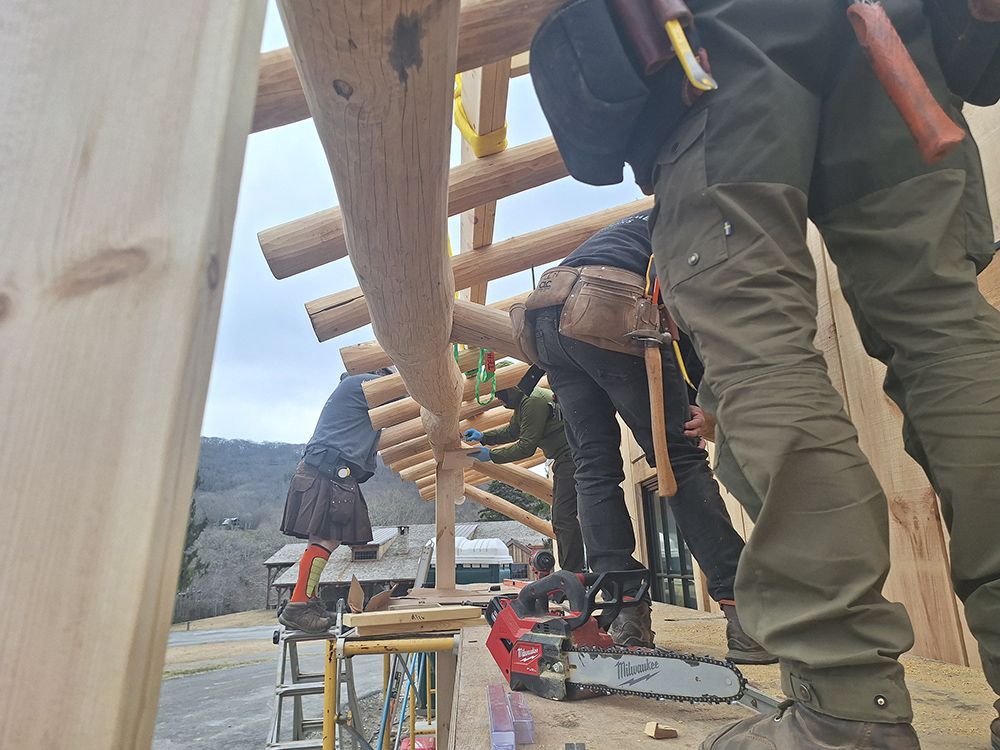 Construction workers building a wooden structure. Logs, tools, and a chainsaw are visible.