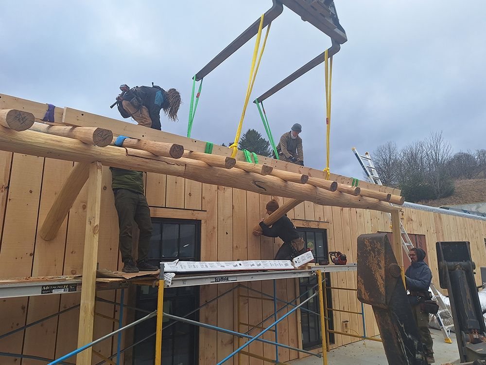Construction workers lifting wooden beams on a building. Scaffolding, crane straps, and sky visible.