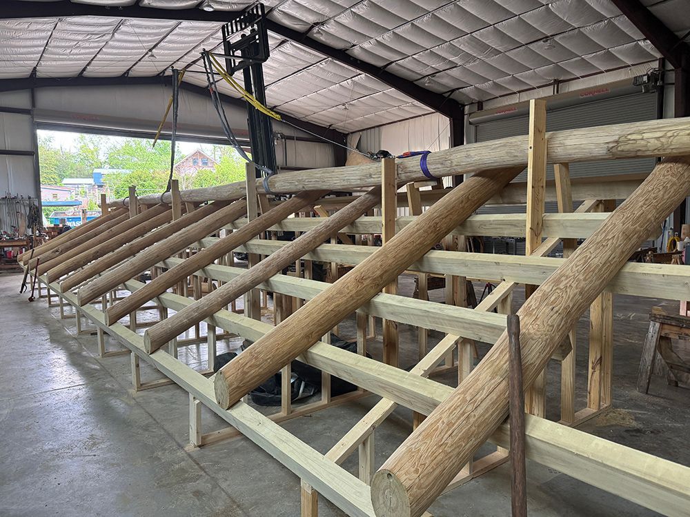 Construction of a wooden structure in a workshop. Logs are being positioned atop a frame, with a person operating machinery.
