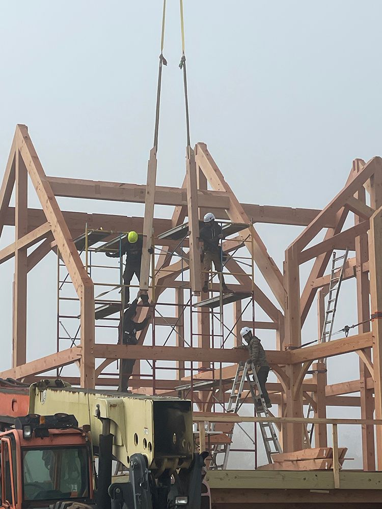 Construction workers assemble a wooden frame building using a crane. Foggy sky.