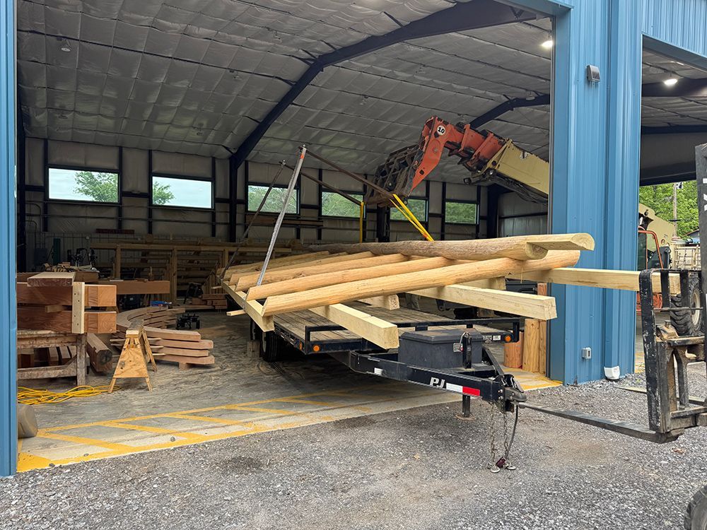 Forklift unloading a trailer loaded with wooden logs inside a blue metal building with an open bay door.
