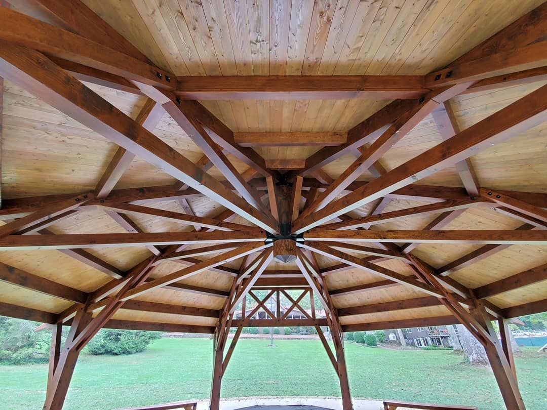Wooden gazebo ceiling with radiating beams, supporting a light-colored wooden roof, viewed from below.