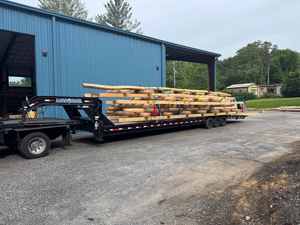 A trailer loaded with logs sits in front of a blue building, on a gravel surface.