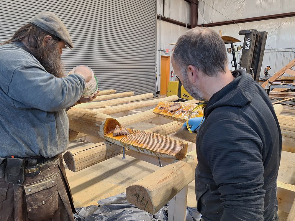 Two people working on a wooden structure, applying sealant. Workshop setting, one with beard, other wearing glasses.
