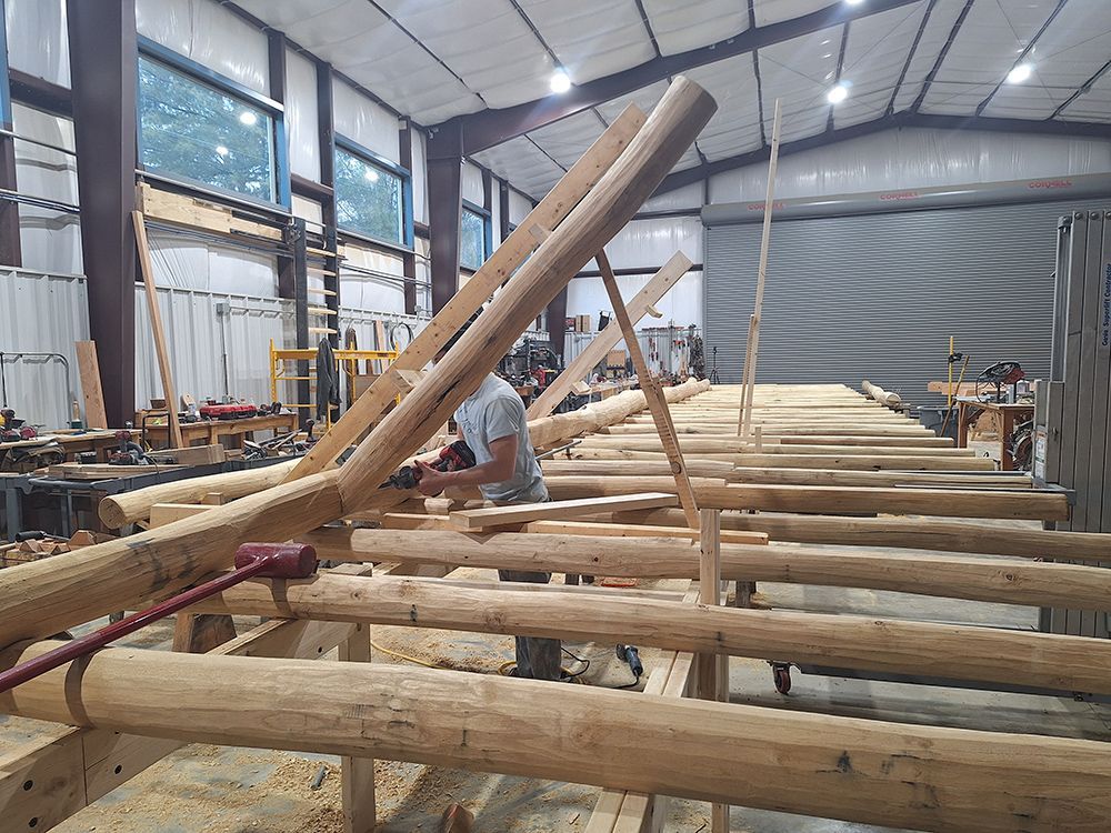 Man working on a log cabin frame inside a metal building, using tools to secure logs.
