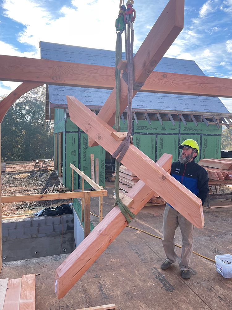 Construction worker guides wooden beams suspended by a crane. Blue sky, unfinished building in the background.