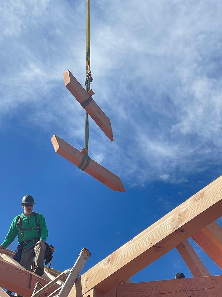 Worker on roof with safety harness watches lumber being lifted by a crane against a blue sky.