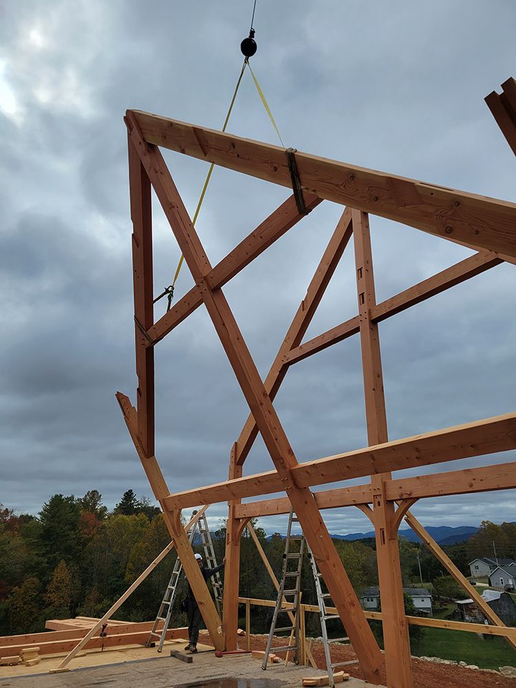 Crane lifting a timber frame structure against a cloudy sky. A worker on a ladder assists.