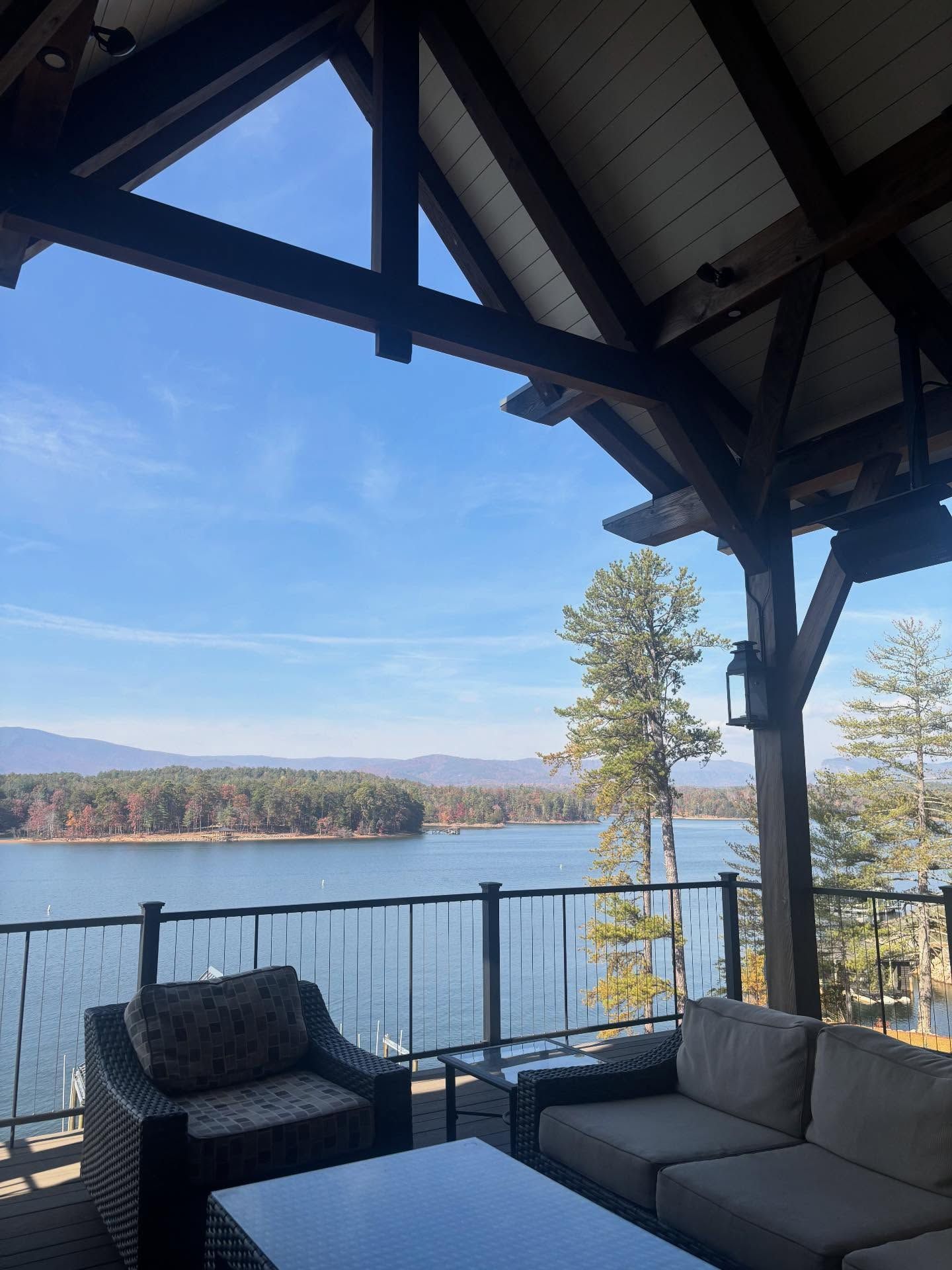 Covered patio overlooking a lake and mountains. Brown wood beams, glass railing, wicker furniture, and blue sky.