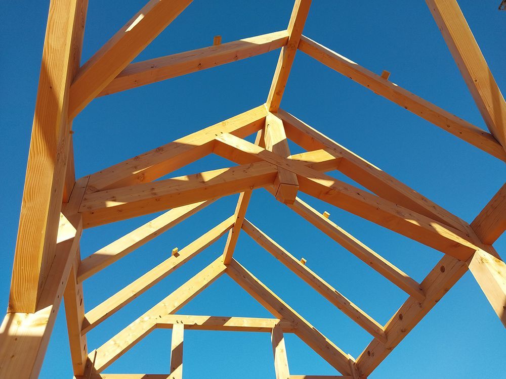 Wooden roof frame against a clear blue sky, showing rafters and a central support structure.
