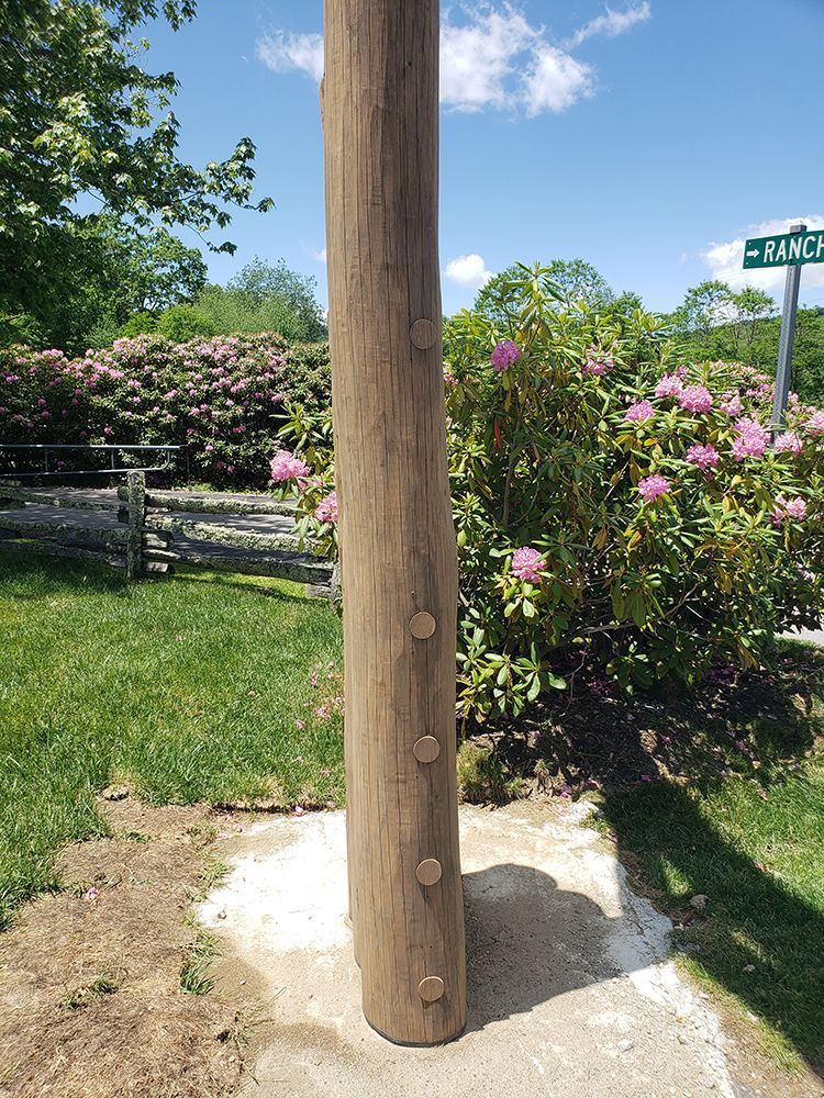Wooden post with embedded pegs on a concrete base, in front of a blooming bush and a street sign.
