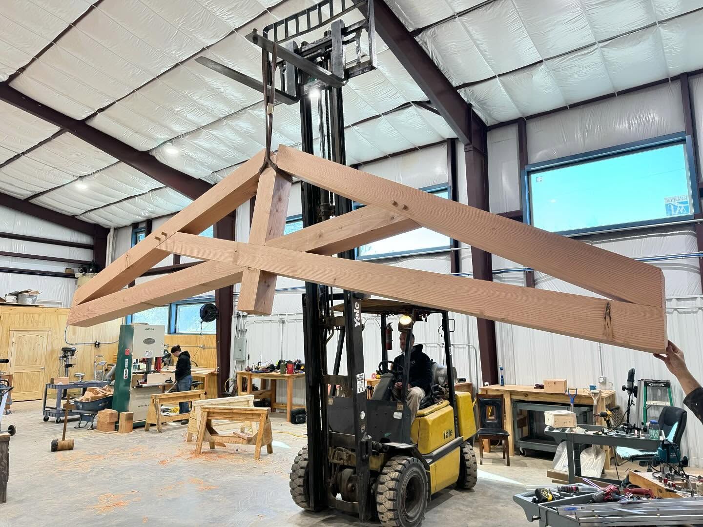 Forklift lifting a large wooden truss inside a workshop with tools and a person visible.