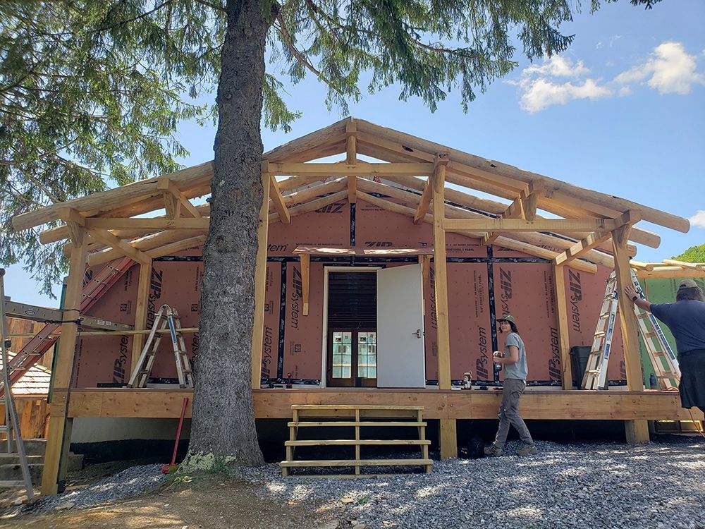 Building exterior with exposed wood frame, red insulation, and a central doorway. Two people are working, ladders present.