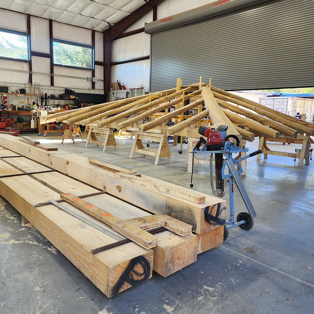Workshop with timber frame roof construction in progress. Lumber stacks, circular saw, and assembly on sawhorses.