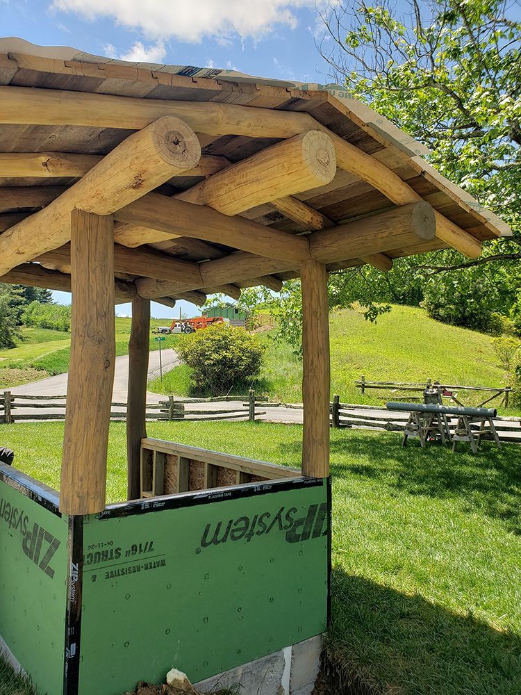 Wooden structure with log beams and posts, green siding, set in a grassy area with trees and a path.