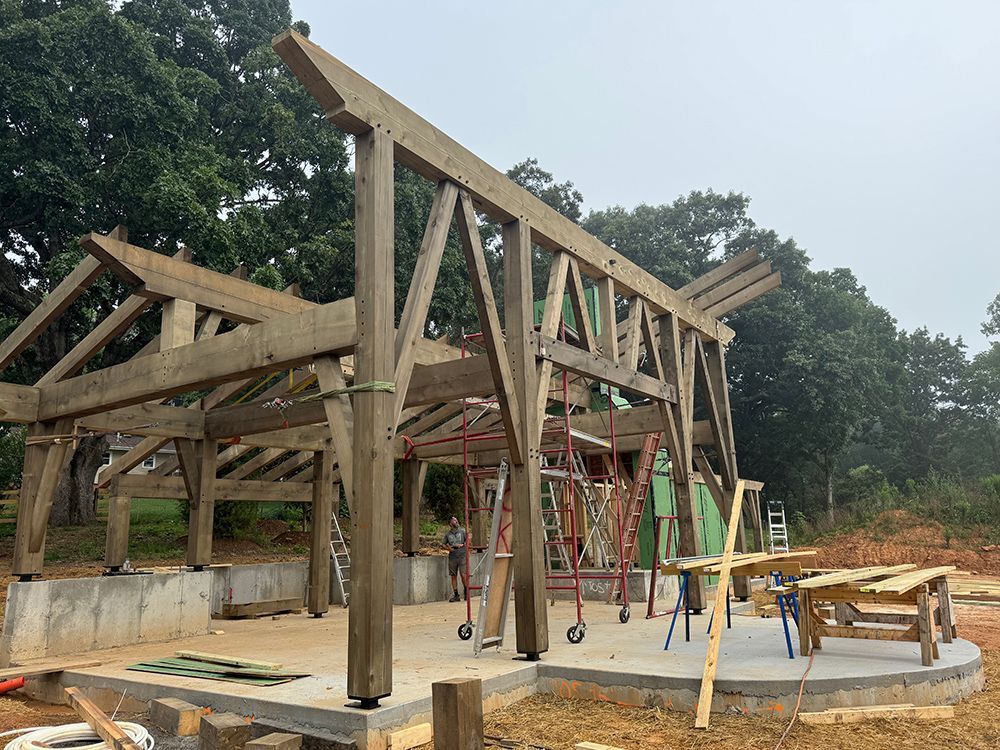 Wooden timber frame building under construction, exterior view. Gray beams, supported by concrete base. Green insulation, scaffolding visible.