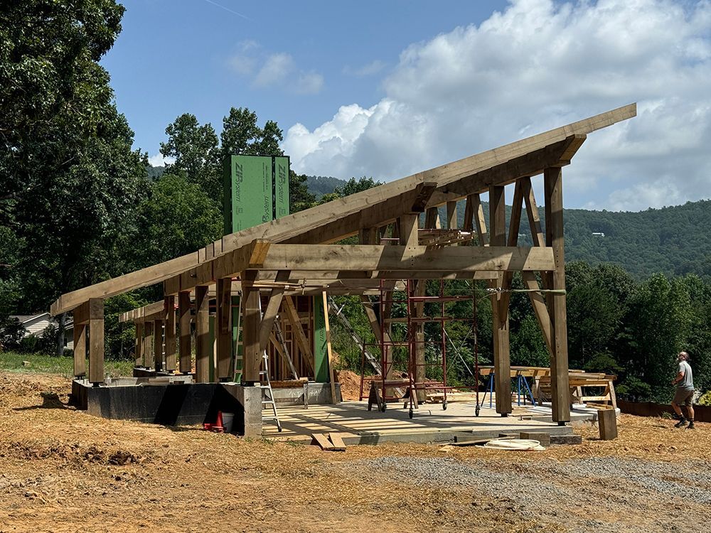 Wooden structure under construction, with angled roof. Man standing nearby on a concrete foundation, mountains in the background.