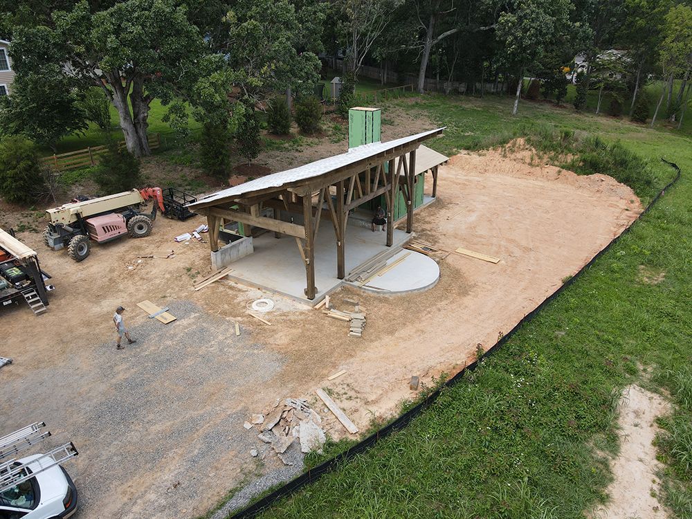 Construction site with a covered pavilion under construction; trees and grass surround the area.