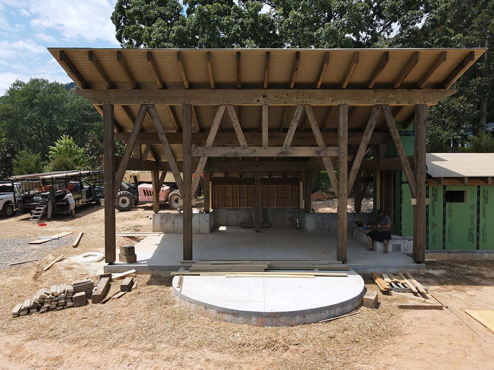 Wooden pavilion under construction, concrete foundation. Construction workers, trees in background.