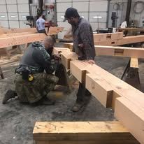 Two men working with large wooden beams in a workshop setting.
