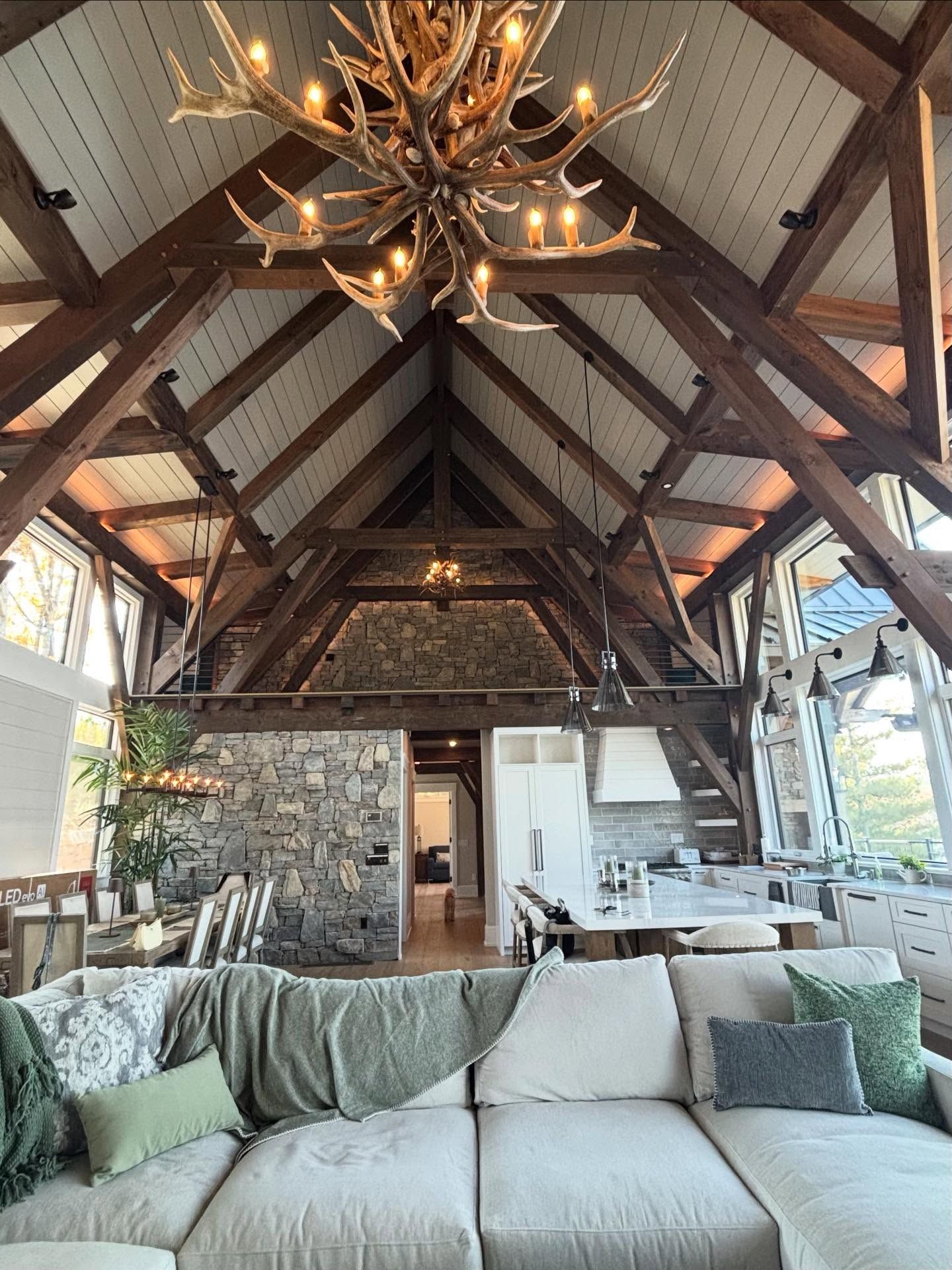 Spacious living room with high wood-beamed ceiling. Features a large antler chandelier, stone wall, and a cream-colored sectional sofa.