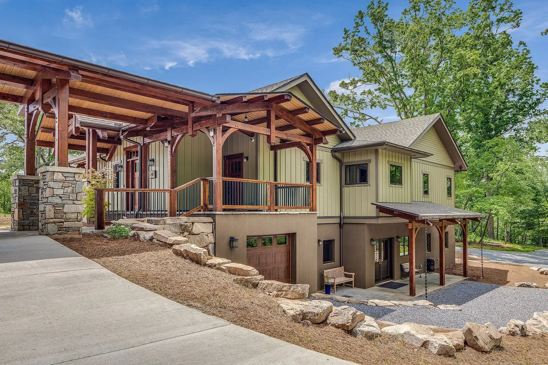 A two-story mountain home with a wooden porch and driveway, nestled in a lush green landscape.