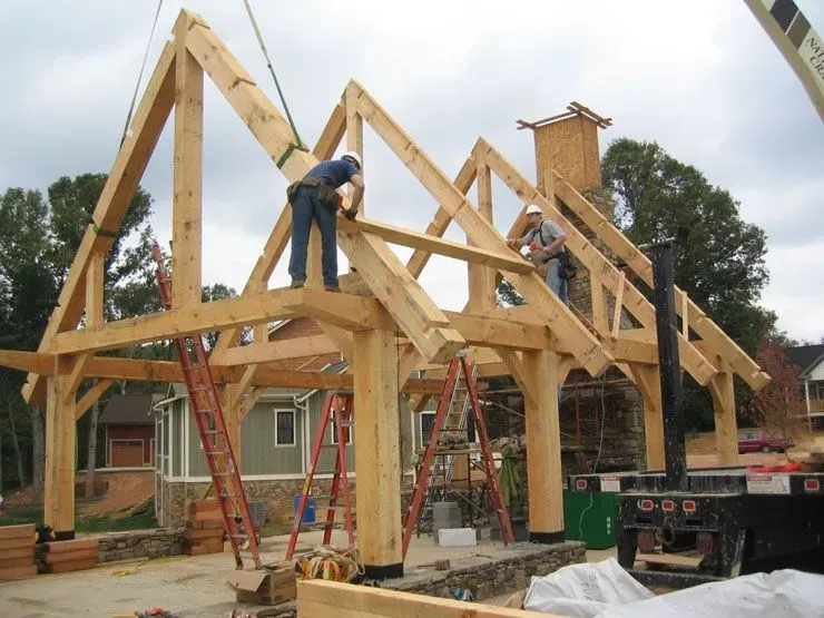 Construction workers assembling a wooden timber frame for a house exterior, with crane, ladders, and building materials.