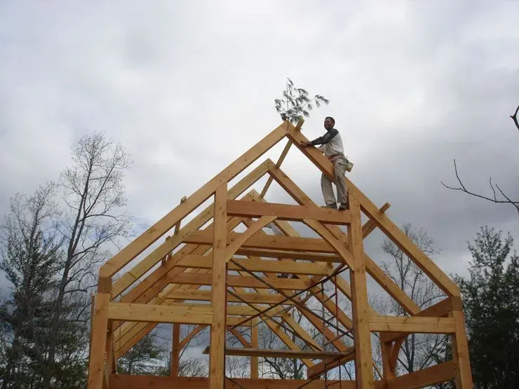 Carpenter atop wooden frame structure under cloudy sky.