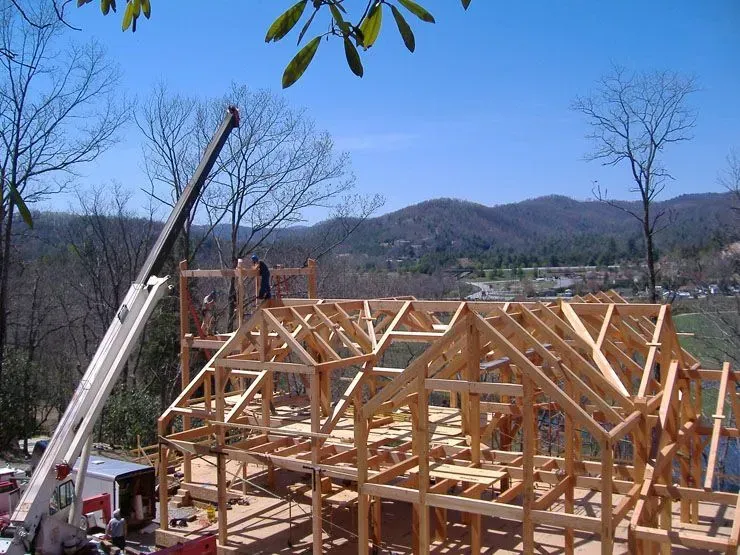 Wooden house frame under construction with crane, sunny day, mountains in the background.