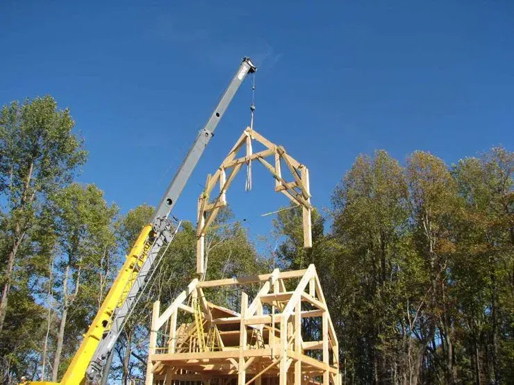 Crane lifting a wooden frame for a house against a clear blue sky, trees in background.