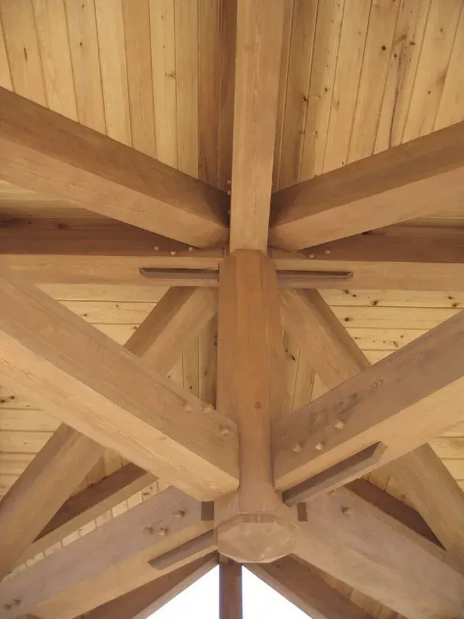 Wooden beams forming a roof structure, joined at a central column. Natural wood tones.