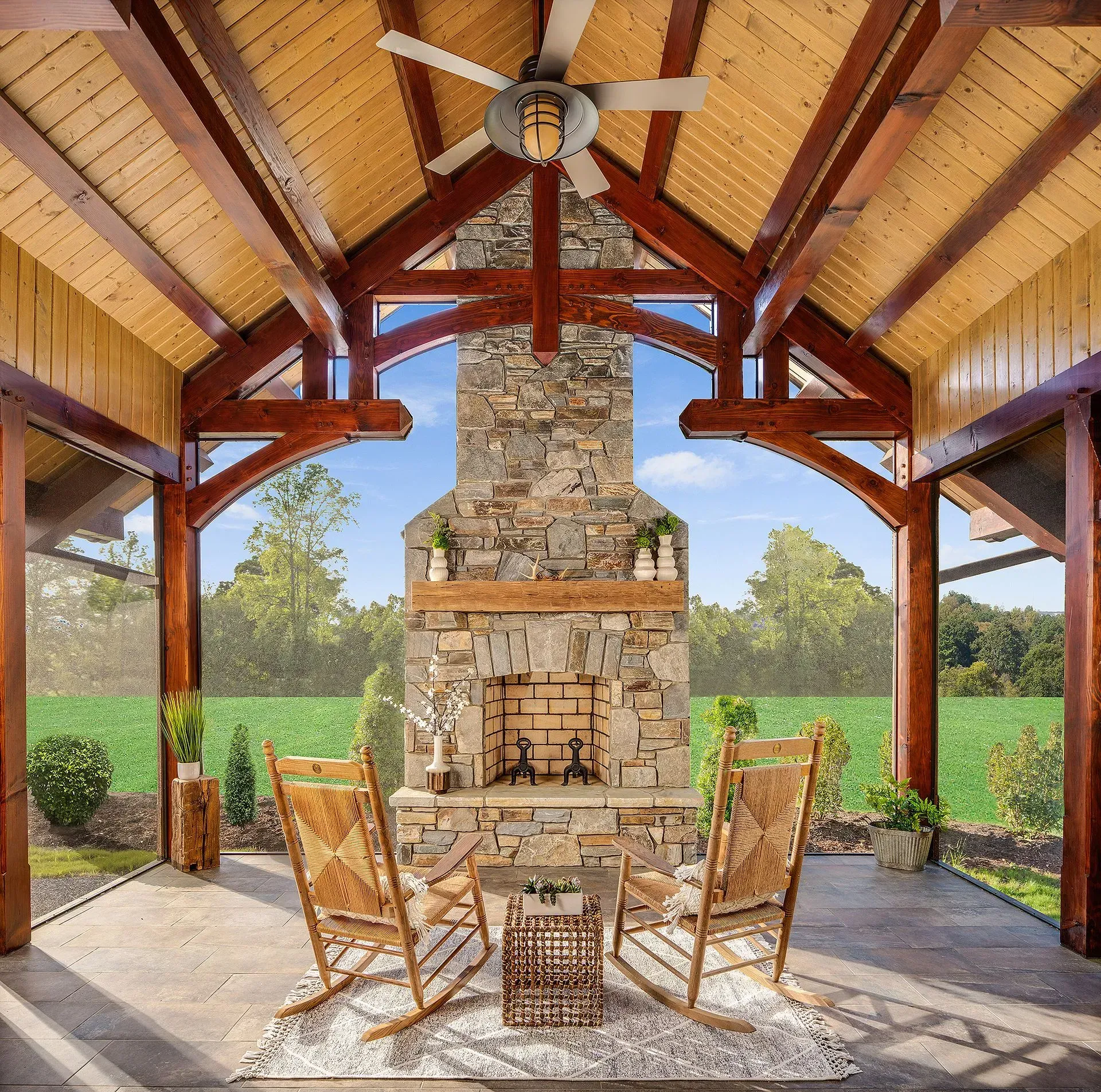 Covered patio with stone fireplace, rocking chairs, and a field in the background.