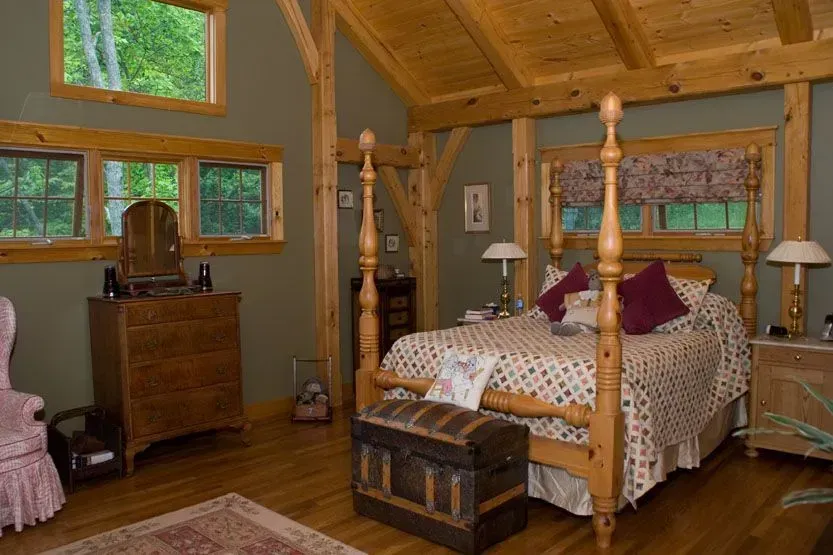 Bedroom with wooden beams, four-poster bed, windows, antique furniture, and a rug.