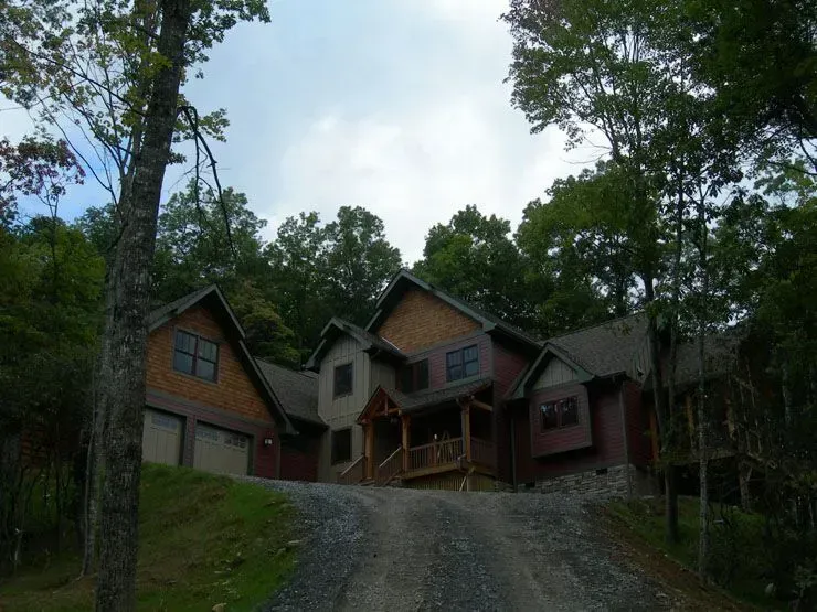 A cabin-style house nestled in a forest, viewed from a gravel driveway.
