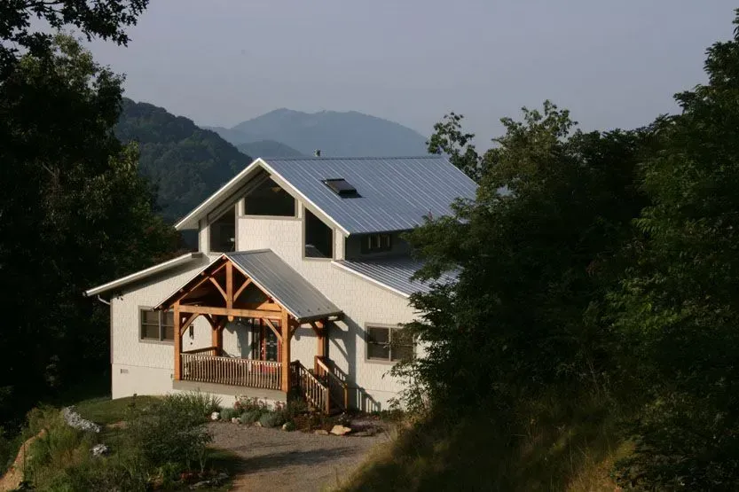 Two-story white house with wooden porch, metal roof, nestled in green trees, mountain backdrop.