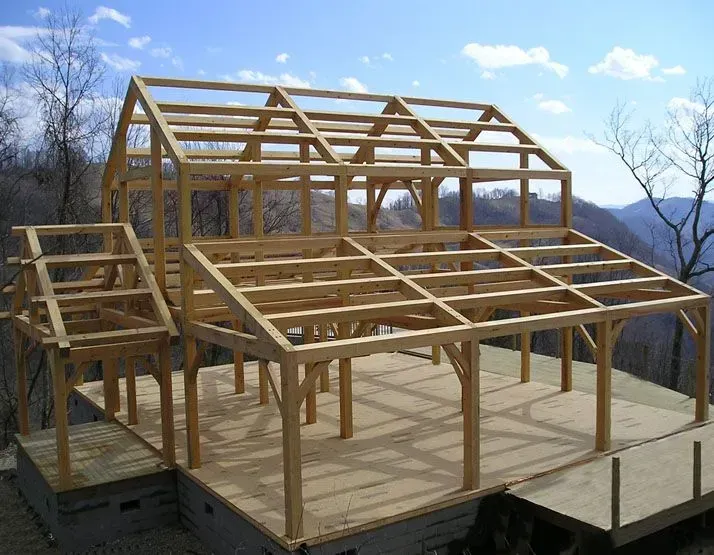 Wooden timber frame of a house under construction on a concrete foundation, set on a hillside.