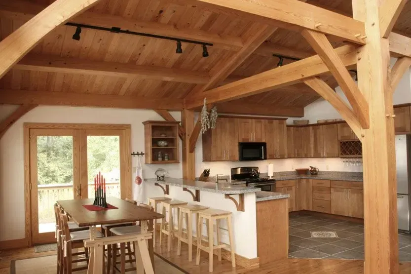 Light-filled kitchen with wooden beams and cabinets, island with stools, and a table near glass doors.