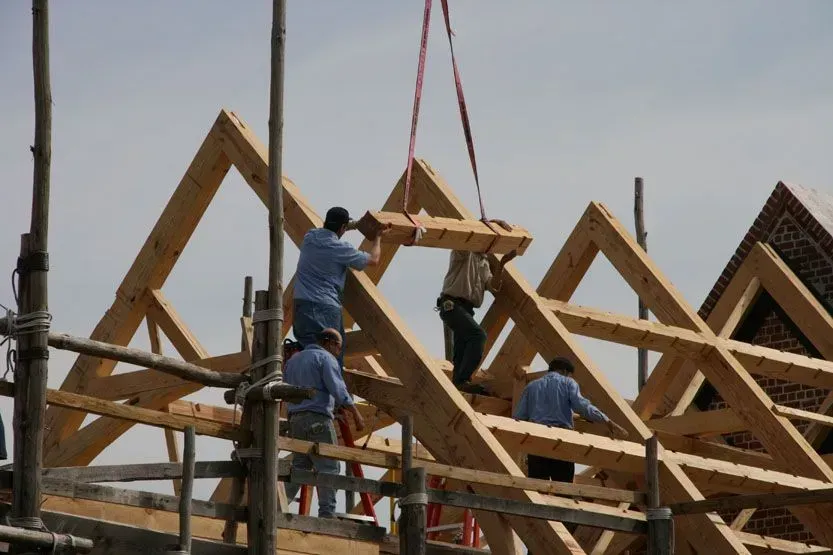 Construction workers placing roof truss pieces on a partially built wooden structure. Sunny day.