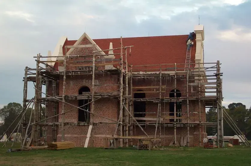 Building under construction with scaffolding; red brick facade, red roof, man on ladder.