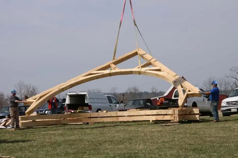 Wooden arch being lifted by a crane during construction, with workers and vehicles in the background.
