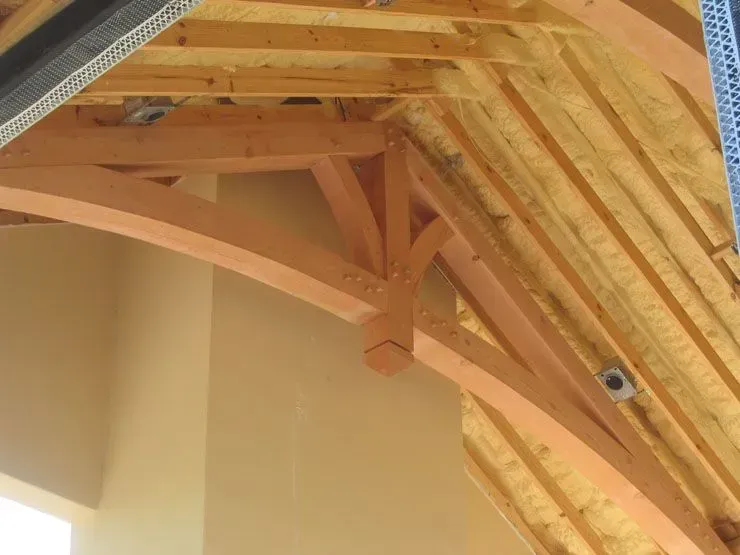 Wooden truss supporting a roof overhang. Light brown beams with complex joinery, set against a pale wall and roof sheathing.