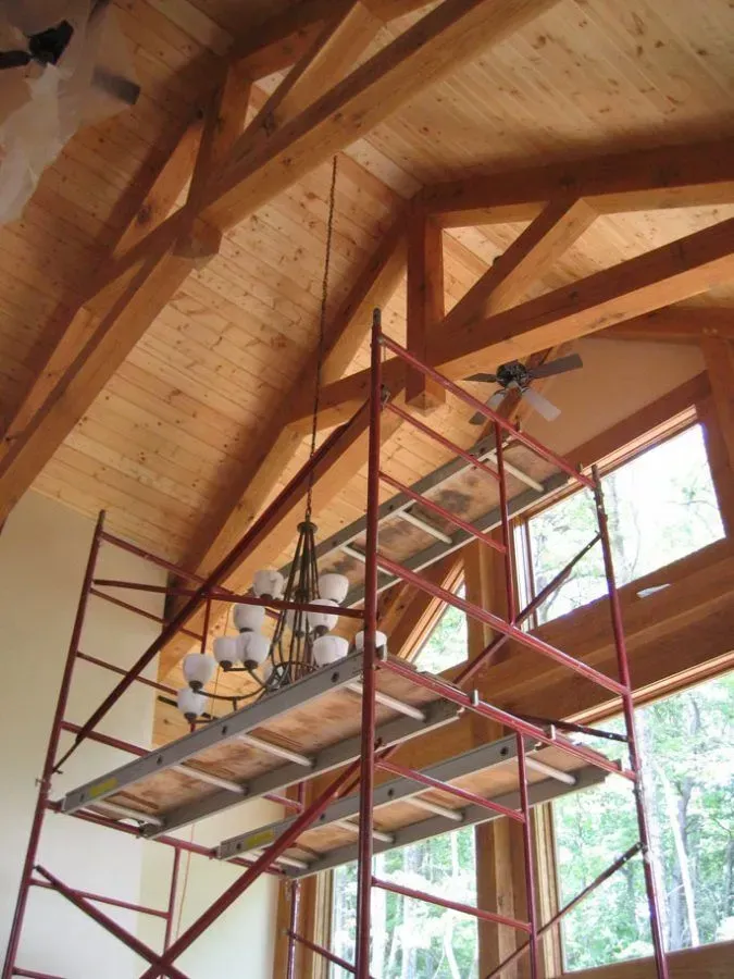 Red scaffolding supports a worker near a window, accessing a chandelier in a room with wood ceiling beams.
