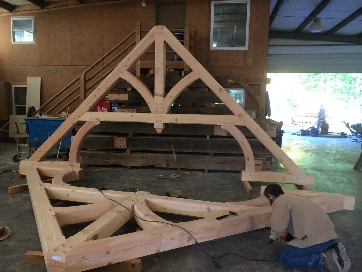 Large wooden framework being assembled by a person kneeling on the ground in a workshop.