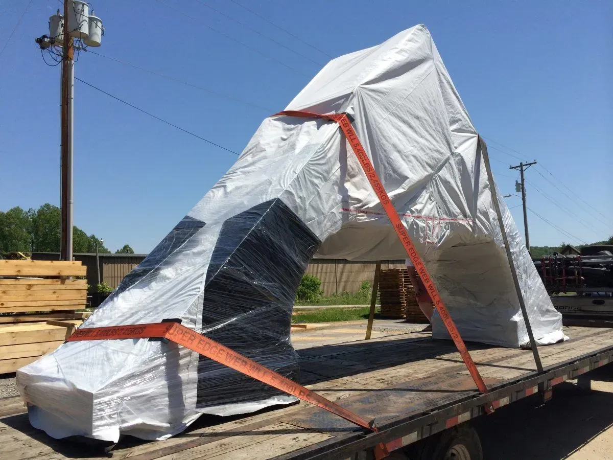 Large, covered object strapped to a flatbed truck on a sunny day. White tarp covers the object, orange straps secure it.