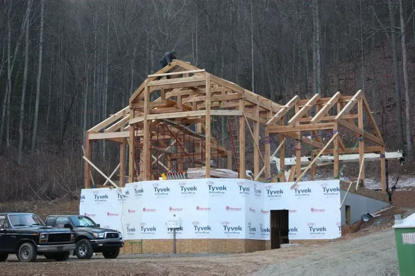 Wooden frame of a house under construction; a worker on the roof; Tyvek wrap on the foundation; vehicles parked nearby, forest in the background.